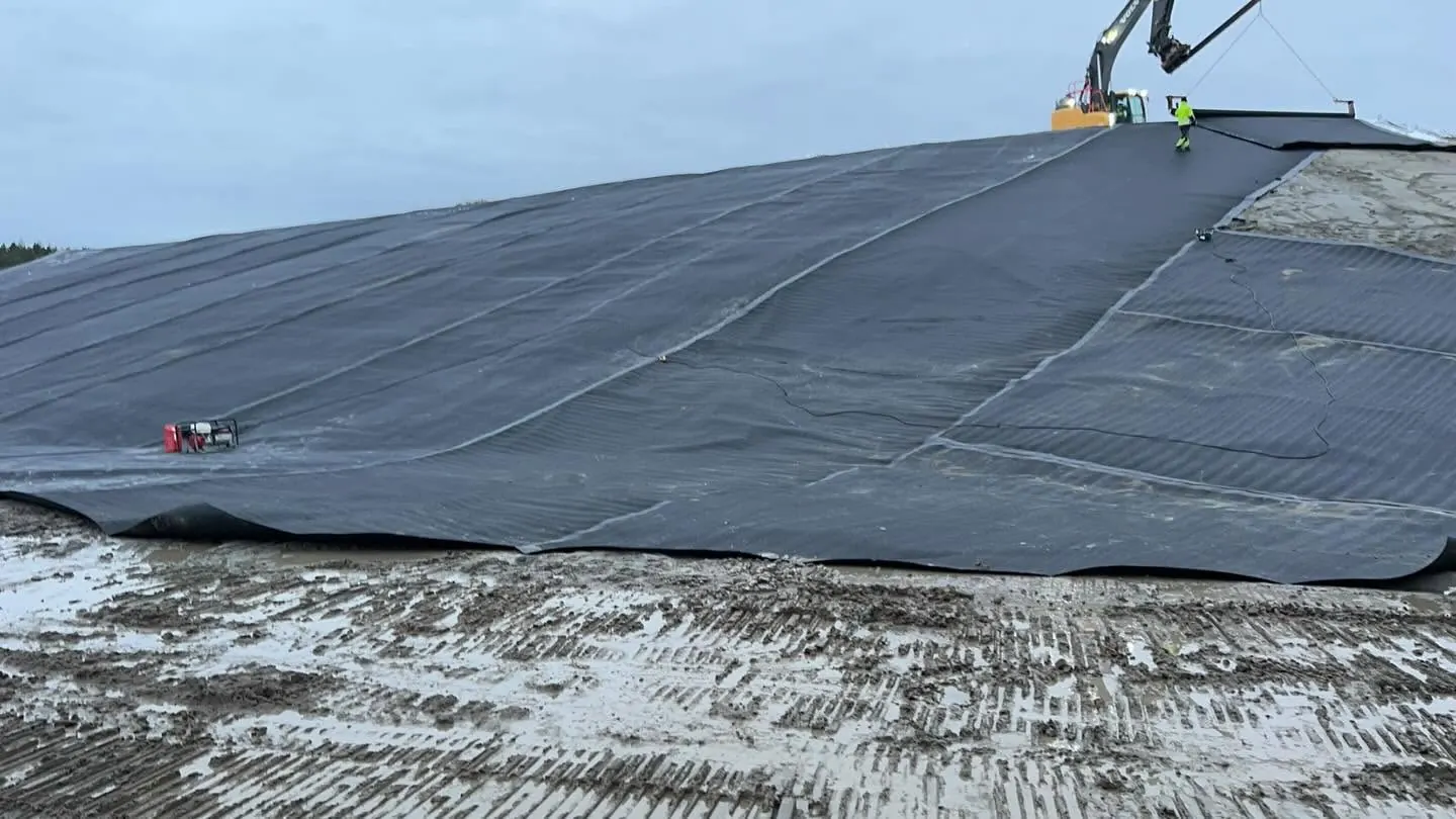 Svart geomembran med svetsade skarvar utlagd på en sluttning, med en grävmaskin och en montör i varselkläder på krönet och ett rött aggregat på membranen