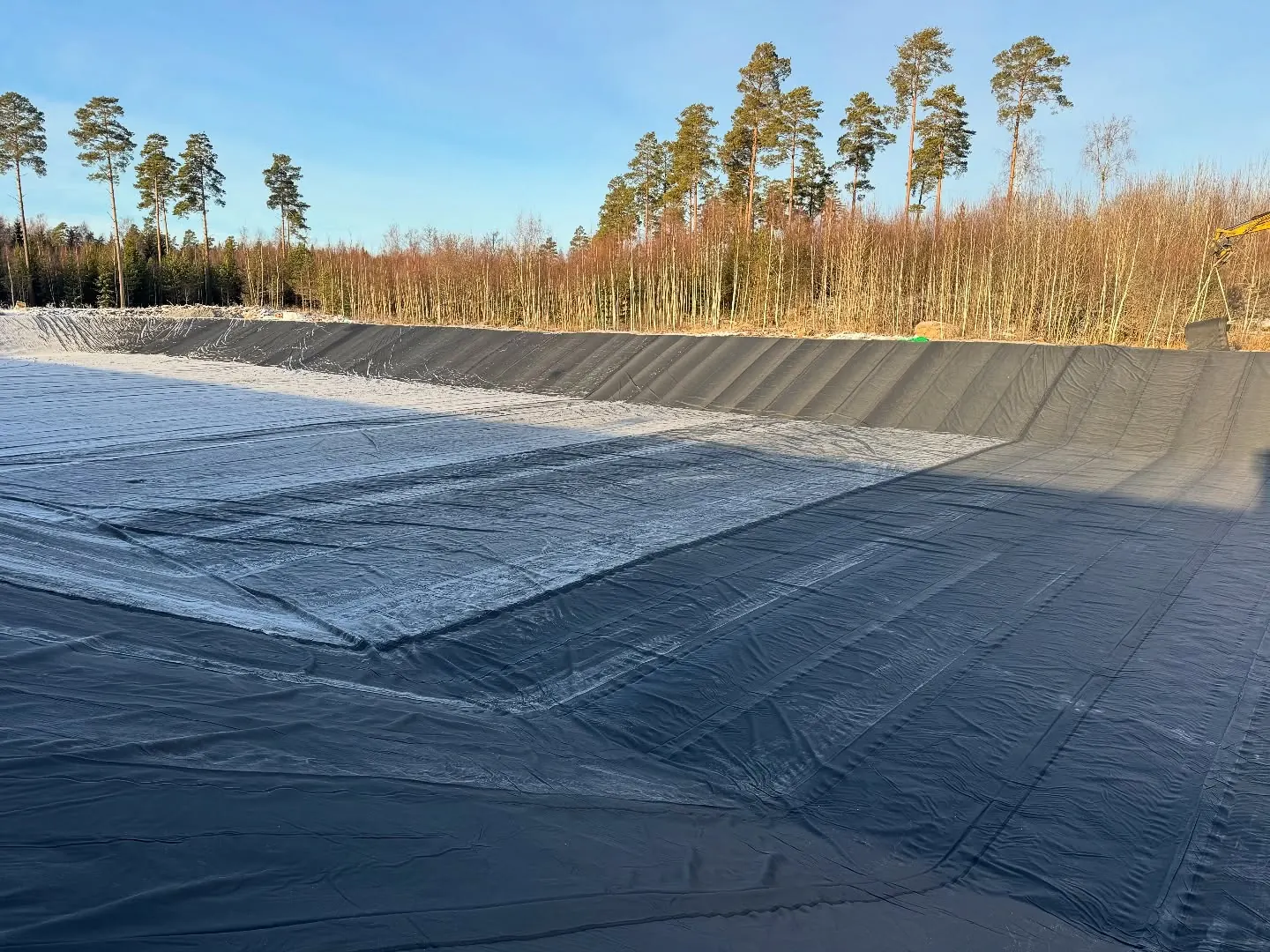 Utgrävd damm klädd med svart geomembran med synliga skarvar, frost i botten och grävmaskin vid kanten, omgiven av tallskog under klar himmel