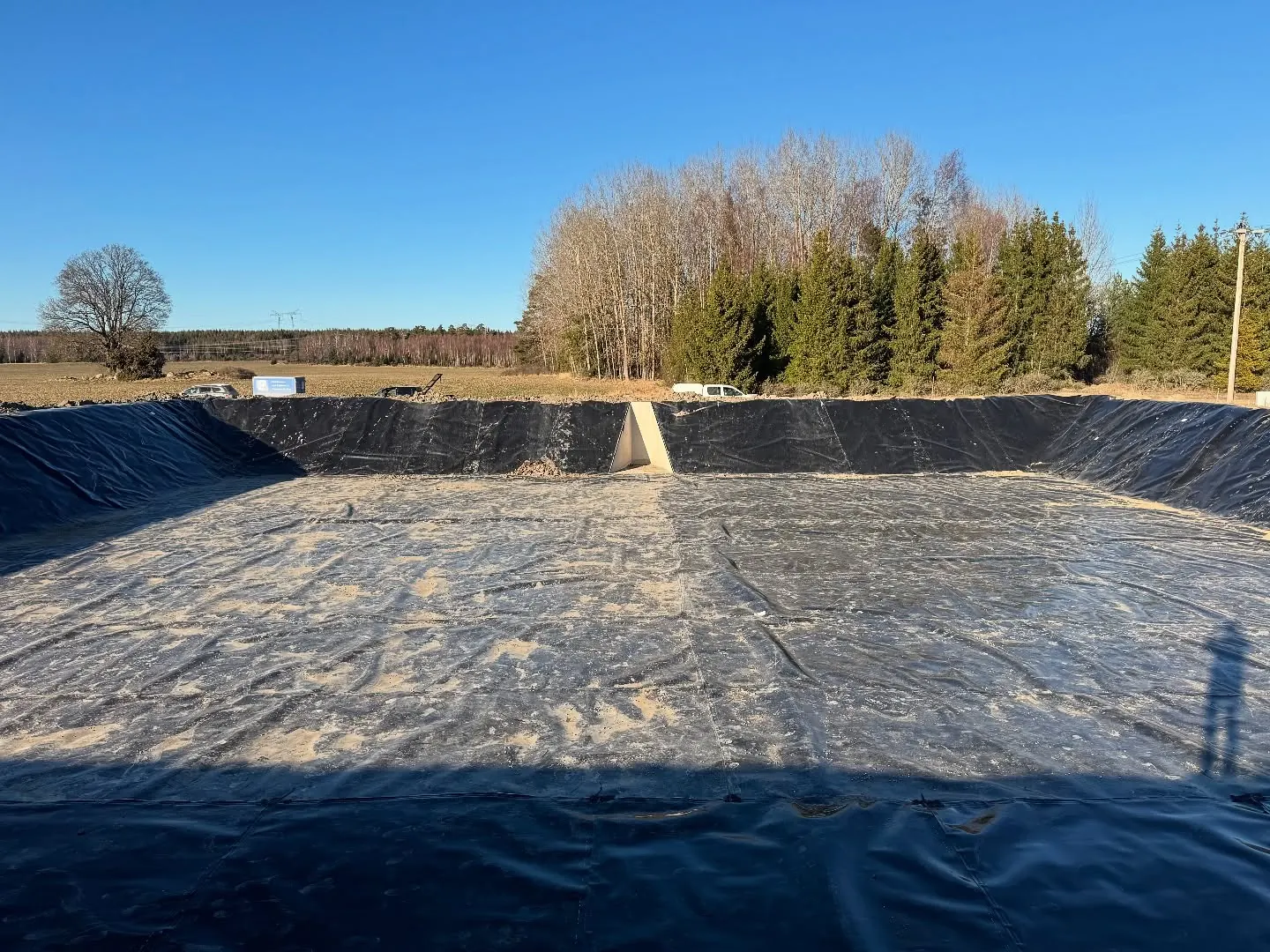 Stor rektangulär bassäng vars botten och sluttande kanter är klädda med svart geomembran med sandfläckar, ljusgrå betongränna i mitten av den bortre vallen, öppna åkrar och en rad av barrträd och lövfria träd i bakgrunden under klar blå himmel
