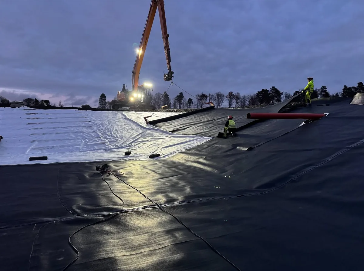 Montörer lägger svart geomembran över vit geotextil i skymningen med lyftkran och grävmaskin i bakgrunden