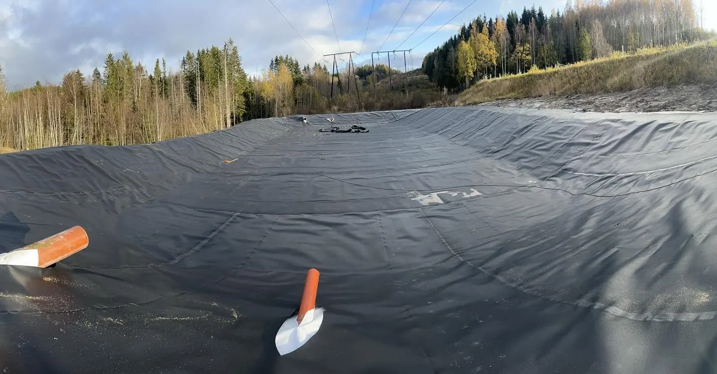 Bassängklädd med svart geomembran med två orange rör och vita packningar i förgrunden, omgiven av jordbankar och tallskog under en högspänningsledning