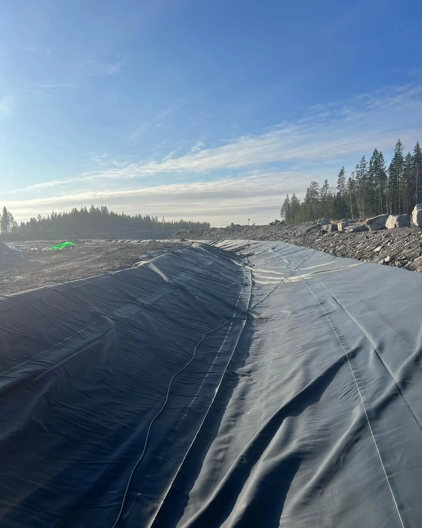 U-formad schaktkanal klädd med veckat svart geomembran med längsgående skarvar, vall av sprängsten och grus till höger och barrskog i bakgrunden under klar himmel